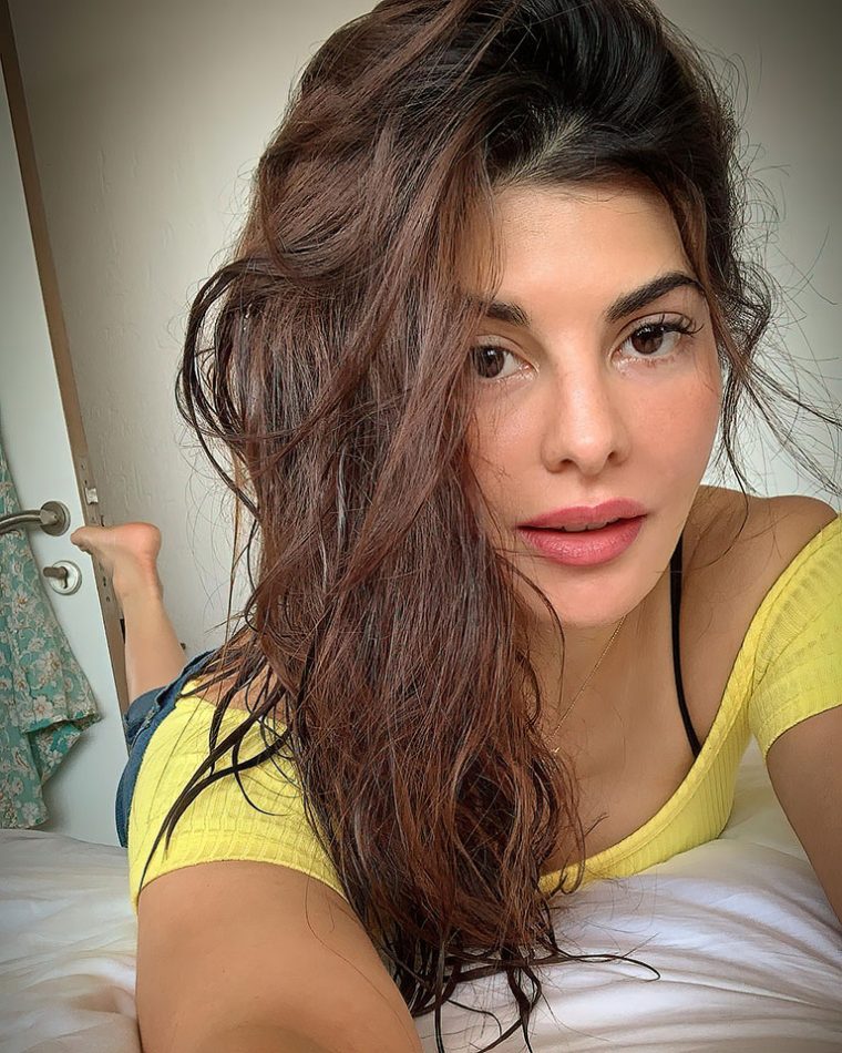 Jacqueline Fernandez laying on the bed wearing a yellow top, black bra and jeans